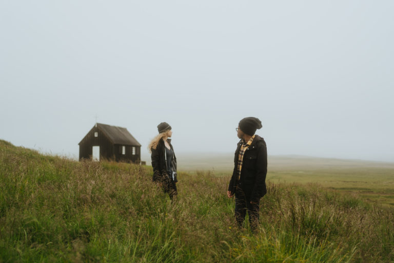 An adventurous couple poses for a photoshoot in Iceland's breathtaking countryside. The pair stand on a lush green grass field. The iconic black church stands tall in the background, adding a striking contrast to the picturesque landscape.